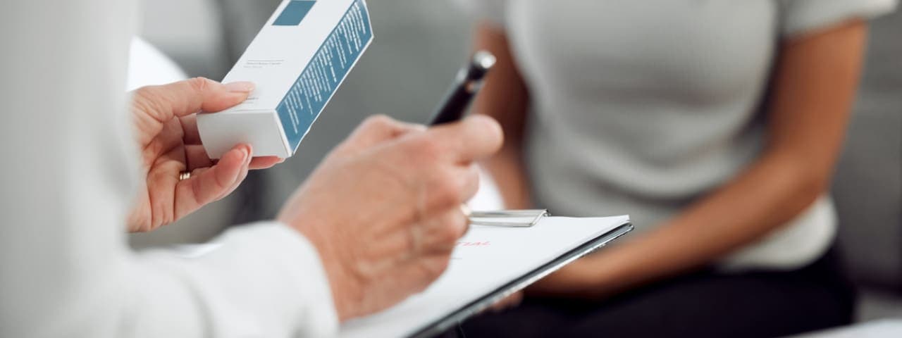 A doctor sitting across from a patient, writing down a prescription for a medication while holding a medication box