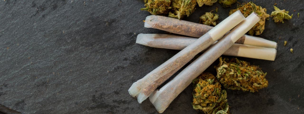 Cannabis joints ready to smoke and a pile of cannabis buds on black stone table.