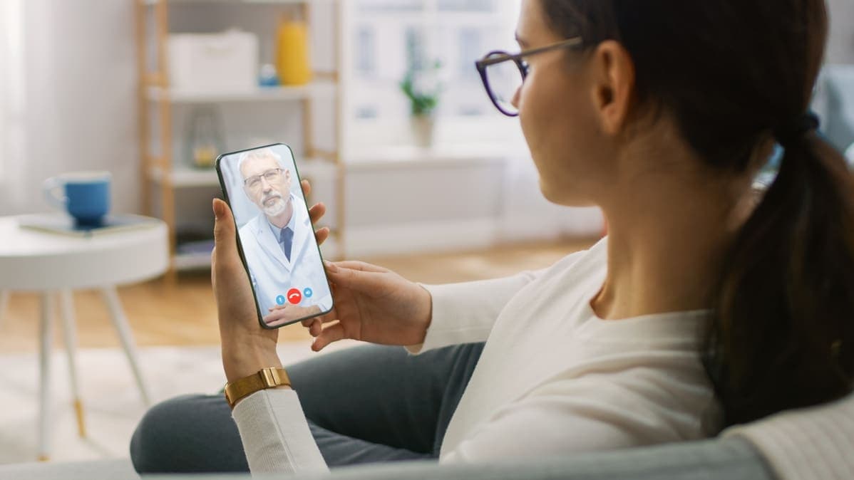 Women sits on a couch in her home speaking with a doctor via a video telehealth call on her smartphone